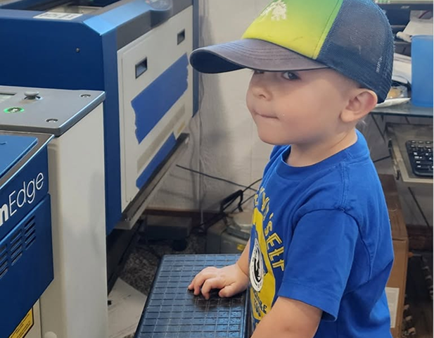 Child standing beside a laser engraving machine at the AW Laser Art workshop
